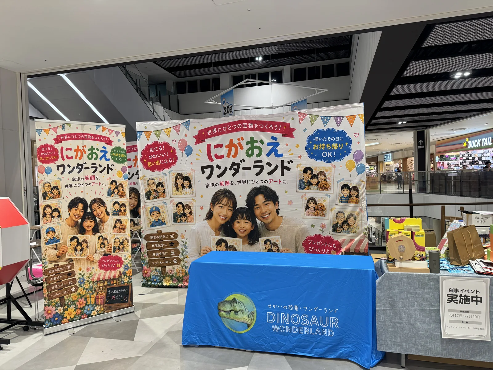 ショッピングモールの通路に設置されたにがおえワンダーランドのブース。大型バナー2本と作品見本パネル、ブランドロゴ入りのテーブルクロスをかけた受付テーブル