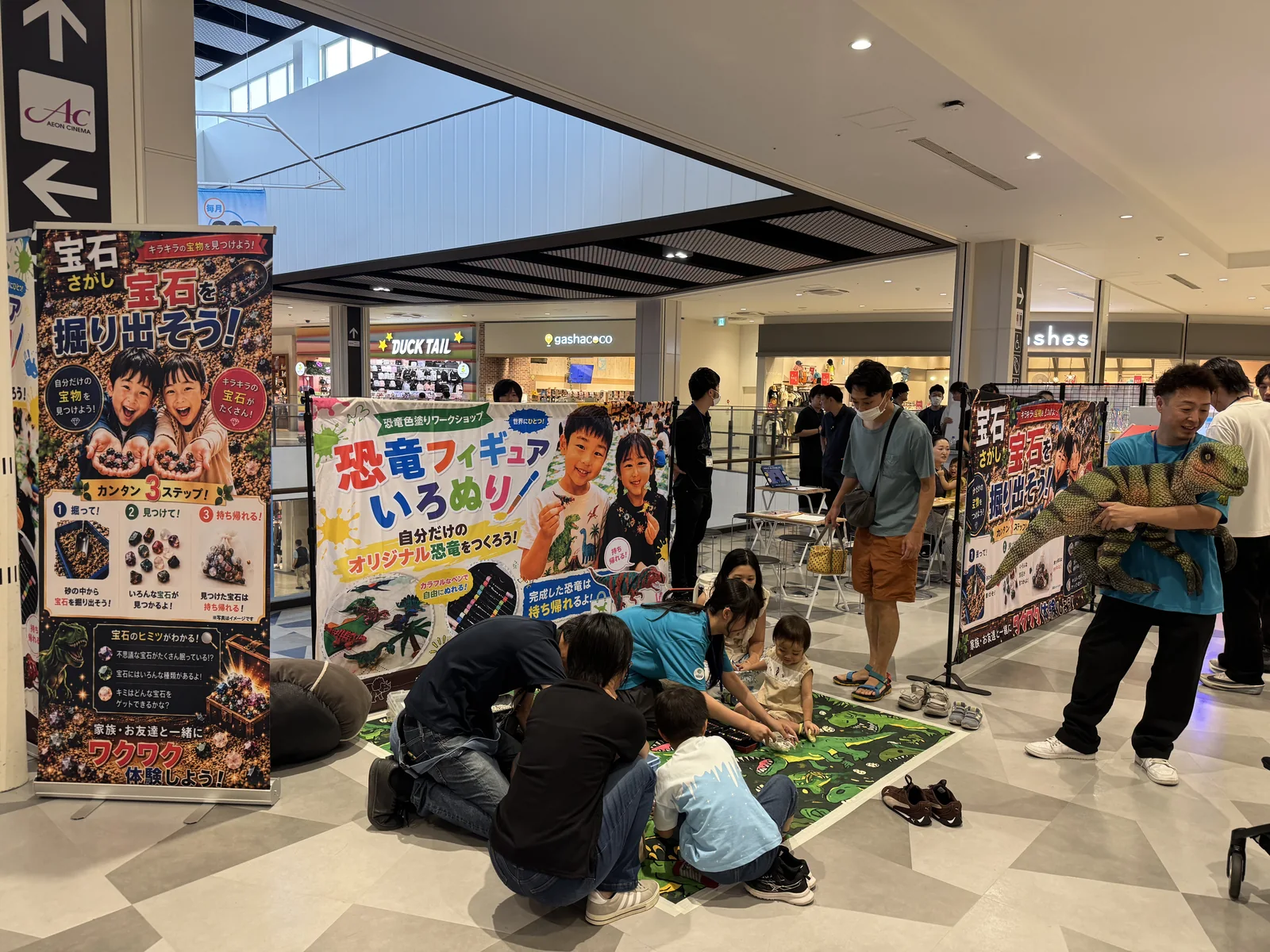 ショッピングモールの通路に設置された恐竜ワークショップのブース全景。宝石さがしと恐竜フィギュアいろぬりの大型バナー、恐竜マットの上で色塗りに取り組む子どもたちと保護者、赤ちゃん恐竜パペットを持つスタッフ